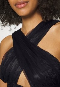 Vestido preto com um design de costas cruzadas em tecido suave e transparente. Camadas texturizadas criam um interesse visual. Área dos ombros exposta.