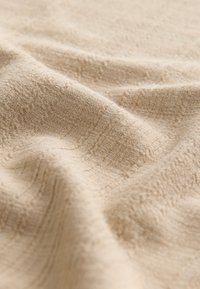 Beige textured fabric with a soft, slightly raised pattern. The material appears smooth and drapes loosely, showing gentle folds.