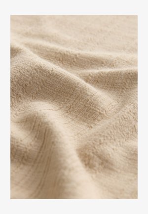 Bēša auduma tekstūra ar mīkstu, nedaudz reljefainu rakstu. Materiāls izskatās gluds un brīvi krīt, veidojot vieglas krokas.