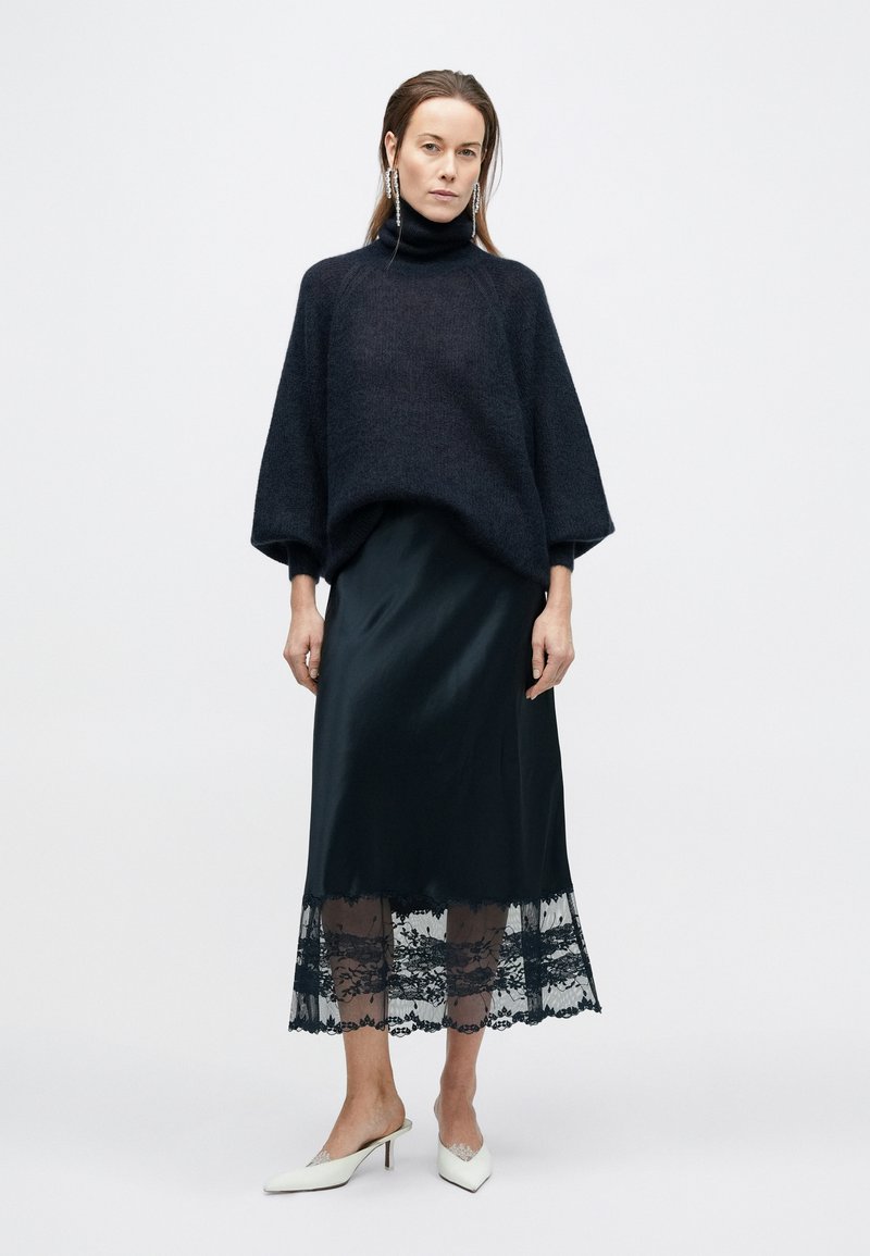 Oversized dark sweater with a high neck; silky black skirt featuring a lace hem. White pointed-toe heels complete the outfit.
