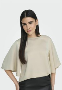 Beige top s volnými, širokými rukávy a kulatým výstřihem, vyrobený z hladkého materiálu, v kombinaci s černou sukní.