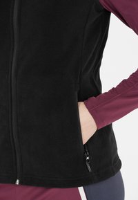 Sort fleece vest med høj krave, lynlås foran og sidelommer. Har en glat tekstur og en kontrasterende bordeaux langærmet skjorte nedenunder.