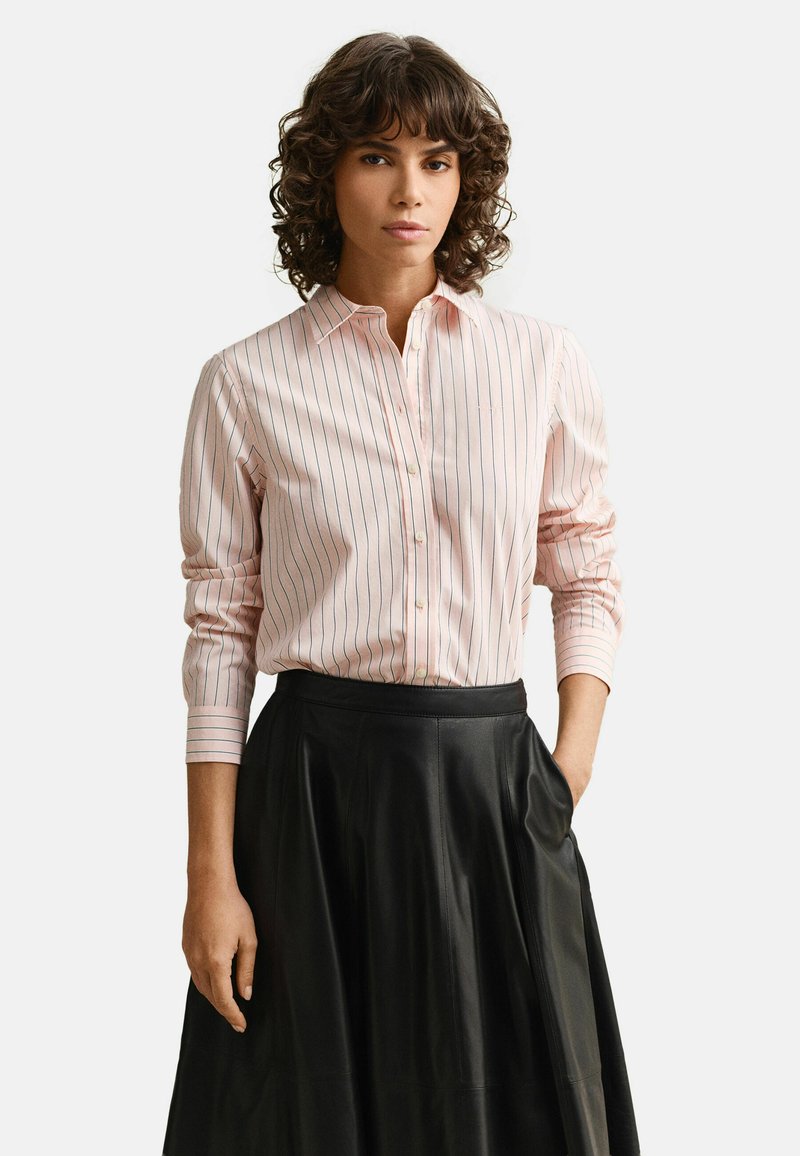 Femme aux cheveux bouclés portant une chemise rayée rose boutonnée, rentrée dans une jupe en cuir noire, debout avec une main dans la poche.