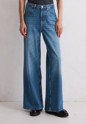 Persoon die een high-waisted wijde blauwe spijkerbroek en zwarte schoenen draagt, staand tegen een effen lichte muur.