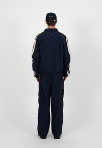 Homme de dos en survêtement bleu marine avec des rayures blanches et jaunes sur les manches et le pantalon, portant une casquette assortie bleu marine, debout sur un fond uni.