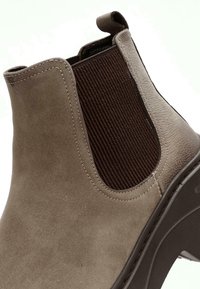 Bottine en daim marron avec des panneaux élastiques, texture lisse, bout rond et semelle en caoutchouc. Dotée d'une languette à l'arrière.