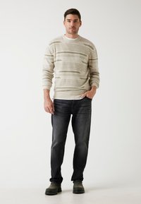 Beżowy sweter z dzianiny w zielone paski, zestawiony z ciemnoniebieskimi dżinsami. Model ma na sobie czarne buty, stoi na jednolitym tle.