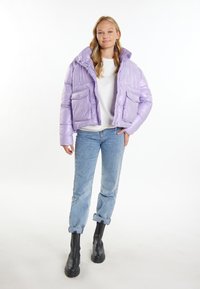 Giacca puffer lavanda con design corto, dotata di due tasche frontali, bottoni a pressione e texture brillante, abbinata a jeans azzurri e stivali neri.