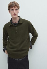 Ciemnozielony sweter z dzianiny z prążkowanym wykończeniem i kołnierzem, wyposażony w trzy guziki i luźny krój, założony na szarym T-shircie.