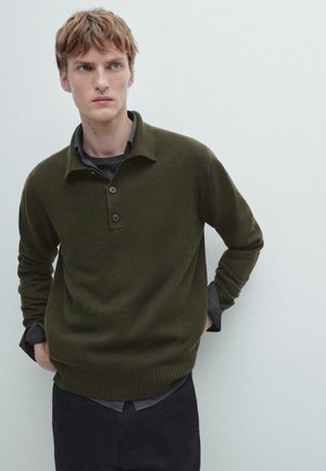 Pullover lavorato a maglia verde scuro con orlo e colletto a coste, dotato di tre bottoni e una vestibilità rilassata, indossato sopra una camicia grigia.
