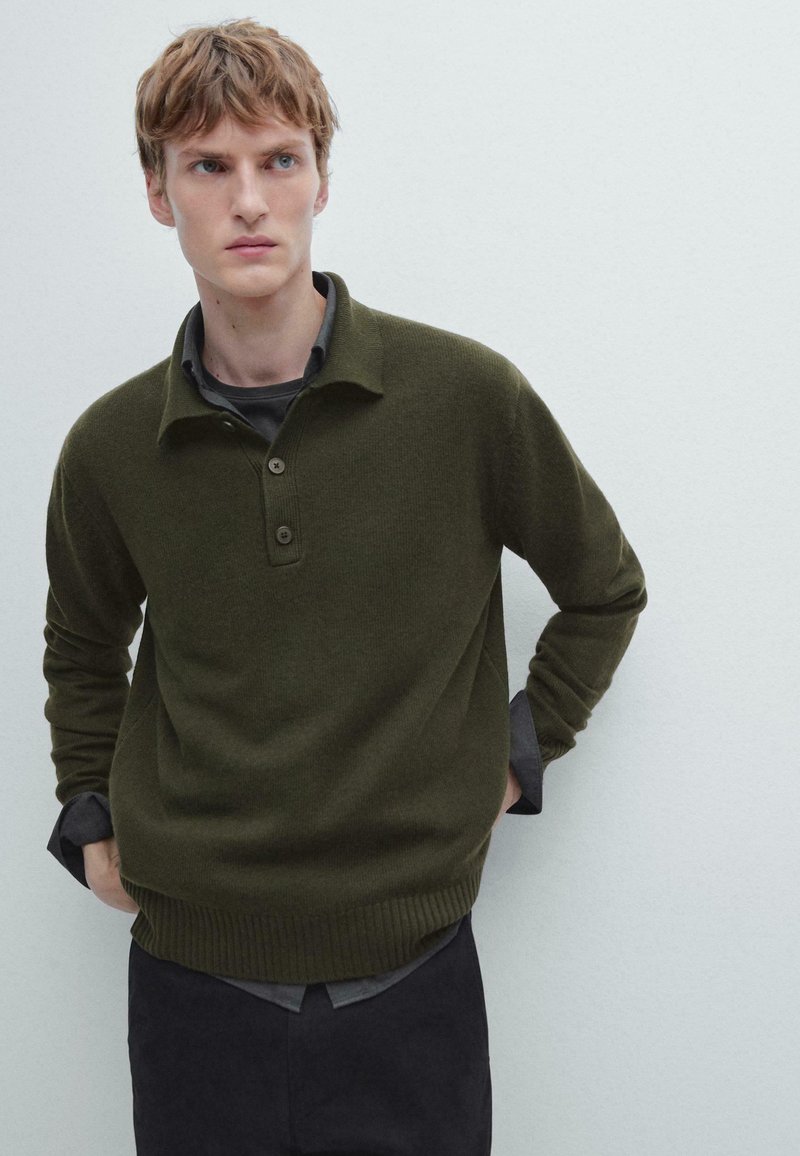 Ciemnozielony sweter z dzianiny z prążkowanym wykończeniem i kołnierzem, wyposażony w trzy guziki i luźny krój, założony na szarym T-shircie.
