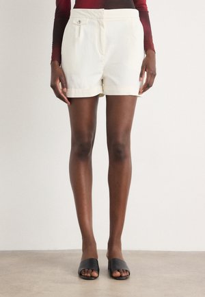 Cremefarbene Shorts aus Baumwolle, mit einem Knopf vorne, seitlichen Taschen und umgekrempelten Säumen, kombiniert mit schwarzen Slide-Sandalen.
