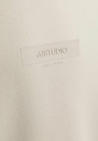 Beiger Baumwollstoff mit einem geprägten Logo-Label "JJ STUDIO" und dem Gründungsjahr "EST. 1990." Die Textur ist glatt und einheitlich.