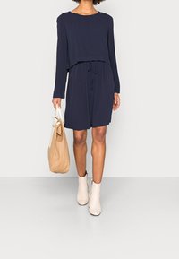 Mujer con un vestido de manga larga azul marino, sosteniendo un bolso tote beige, con botas blancas hasta el tobillo, de pie frente a un fondo claro y liso.