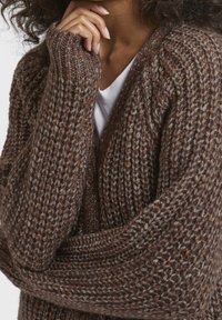 Femme portant un gros cardigan en tricot marron sur une chemise blanche, la main posée près du menton et les cheveux bouclés visibles.