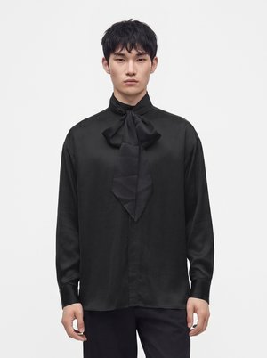 Jeune homme portant une chemise noire en satin à manches longues avec un grand nœud noué au col, debout devant un fond blanc uni.