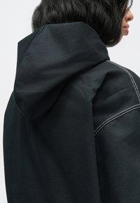 Μαύρο μπουφάν με κουκούλα από υφή ύφασμα, με λευκές ραφές και χαλαρή σιλουέτα, με μεγάλη κουκούλα και λεπτομέρεια στη ραφή των ώμων.