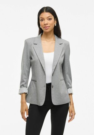 Femme aux longs cheveux noirs portant des boucles d'oreilles en argent, un blazer gris avec les manches retroussées, un haut blanc et un pantalon noir, se tenant contre un fond blanc.