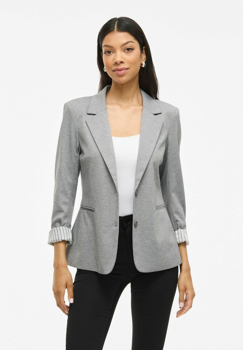 Donna con lunghi capelli scuri, che indossa orecchini a cerchio argentati, un blazer grigio con le maniche arrotolate, una maglietta bianca e pantaloni neri, in piedi su uno sfondo bianco.