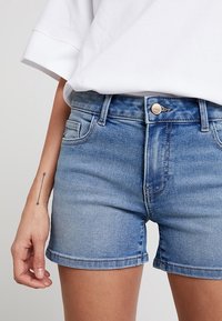 Shorts de mezclilla en lavado azul claro, con un corte recto, cinco bolsillos, un cierre de botón metálico y detalles de costura visibles.