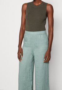 Osoba w ciemnozielonym, bezrękawnikowym topie o prążkowanej strukturze oraz jasnoniebieskich, szerokich spodniach z elastycznym paskiem w talii, stojąca na jednolitym jasnym tle.