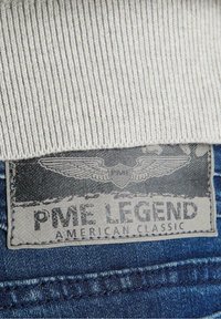 Tela de canalé beige en la parte superior; mezclilla azul oscuro abajo con un parche de cuero gris. El parche presenta texto en relieve y diseño de alas.