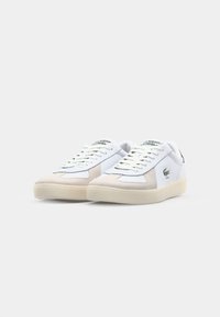 White and beige sneakers with a suede toe cap, featuring green logo accents, flat laces, and a rubber sole. Classic athletic design.