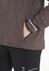 Brun lättviktsjacka med reflektiva detaljer, fickor med dragkedjor och gatherade ärmslut, ihop med svarta leggings med en vit logotyp och randigt mönster.