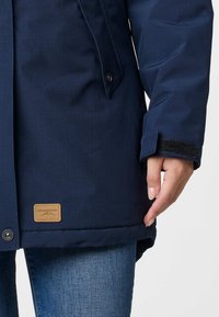 Granatowa kurtka wykonana z gładkiego, wodoodpornego materiału. Posiada naszywkę z logo, zapięcie na zatrzask oraz rzepy na mankietach.