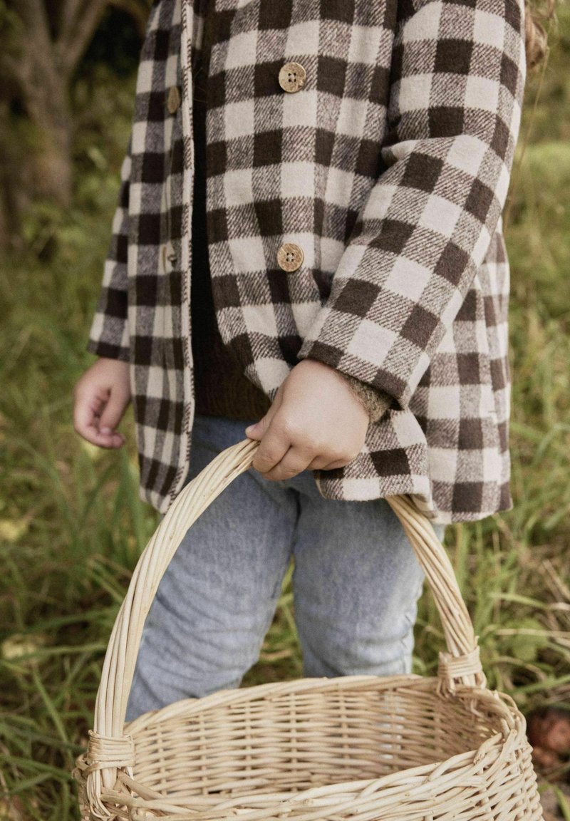 Bambino che indossa un cappotto a quadri marrone e jeans, mentre tiene un cesto di vimini vuoto all'aperto in un'area erbosa.