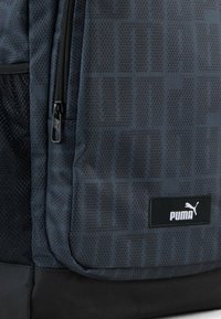 Tamsiai pilkas Puma kuprinė su priekiniais užtrauktukais kišenėmis, tinklinėmis šoninėmis kišenėmis ir Puma logotipu apatinėje priekinėje dalyje.