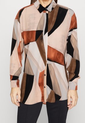 Femme portant une chemise boutonnée à manches longues avec des motifs géométriques abstraits marron, beige, noir et rouille, associée à un pantalon foncé.