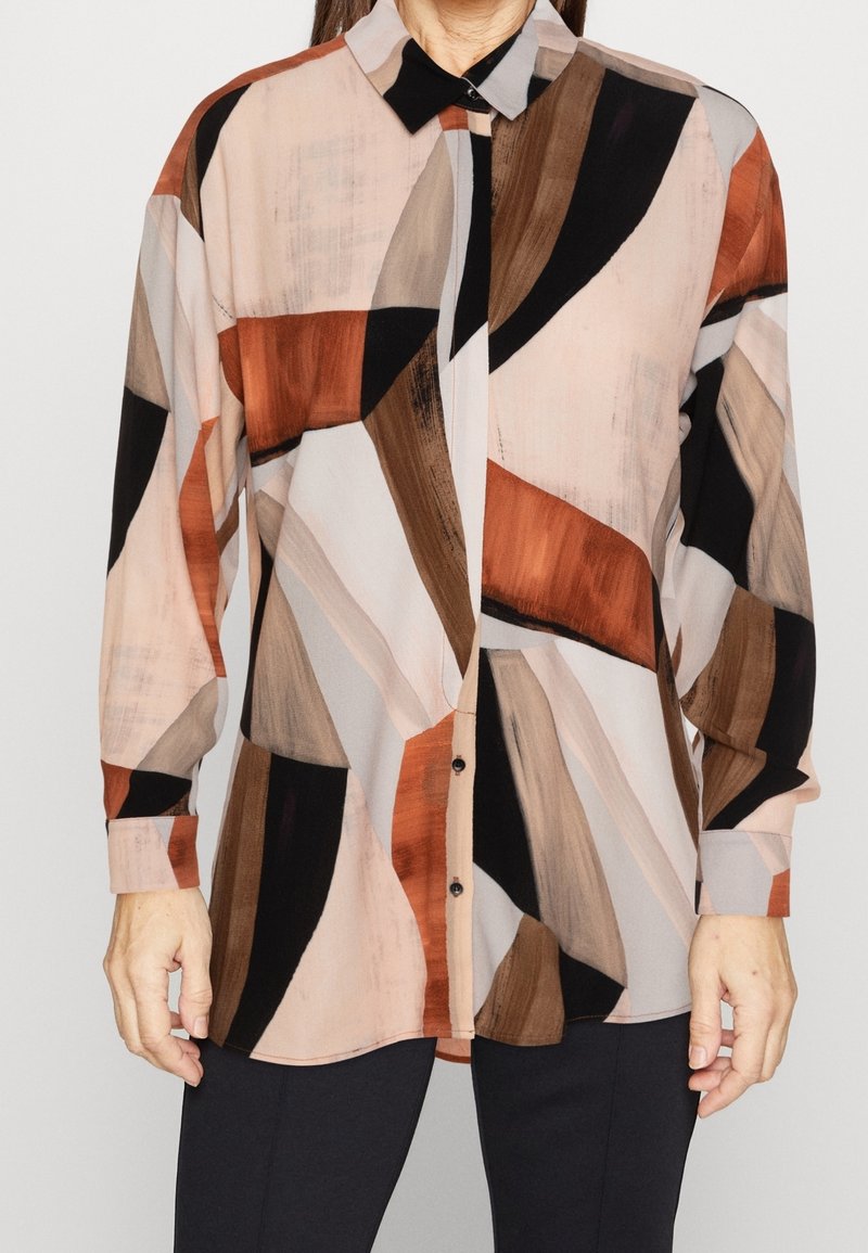 Femme portant une chemise boutonnée à manches longues avec des motifs géométriques abstraits marron, beige, noir et rouille, associée à un pantalon foncé.