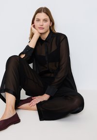 Giovane donna con capelli biondi è seduta sul pavimento contro una parete bianca, indossando una blusa nera trasparente e pantaloni con scarpe ballerine di un viola scuro.