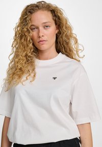 T-shirt bianco in cotone con maniche corte, scollo rotondo e un piccolo logo nero sul petto. Tessuto morbido, vestibilità rilassata.