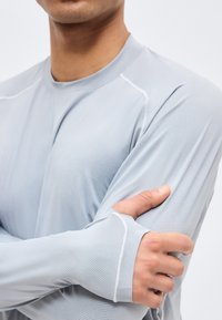 Άνδρας που φοράει αθλητική μπλούζα μακριού μανικιού ανοιχτού γκρι χρώματος με τρύπες για τα αντίχειρα, σταυρώνει τα χέρια του και κρατάει το αριστερό του μπράτσο πάνω από ένα απλό φόντο.