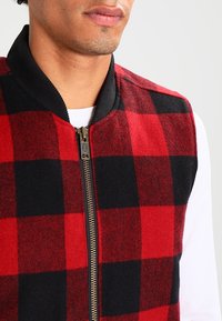 Man med en röd och svart rutmönstrad väst med svart krage över en vit skjorta, visad från halsen till mitten av torso.