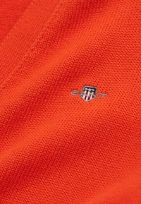Knitted orange fabric with a textured pattern. Features a small embroidered shield logo in gray and black on the lower left.