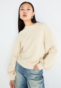 Beige oversized sweatshirt med puffede ærmer og ribbede manchetter, parret med lys blå højtaljede jeans. Enkel, blød tekstur.