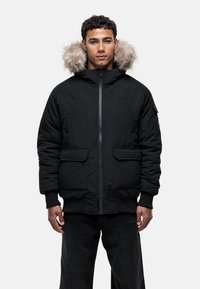 Giacca imbottita nera con cappuccio foderato in pelliccia, due tasche frontali e polsini a costine. Il design è sobrio e funzionale.