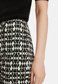 Schwarz-weiße gemusterte Hose mit einem strukturierten Design von Herzformen. Ein kleines Logo ist in der Nähe des Bunds sichtbar.