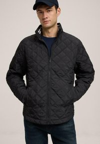 Czarna pikowana kurtka z wysokim kołnierzem i zamek błyskawicznym. Teksturowany wzór o gładkim wykończeniu. Noszona na granatowej koszuli, w zestawie z niebieskimi dżinsami.