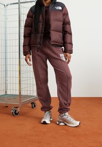 Veste matelassée marron avec accents noirs, pantalon de survêtement bordeaux avec logo, et chaussures de sport gris clair. En arrière-plan, un chariot en métal.