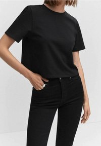 Nainen, jolla on päällä yksinkertainen musta lyhythihainen T-paita ja korkeavyötäröiset mustat farkut. Hänellä on yksi käsi taskussa ja sormus sormessaan.