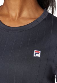 Ναυτικό μπλε αθλητικό μπλουζάκι κατασκευασμένο από υφαντό ύφασμα, με κάθετες ρίγες και ετικέτα λογότυπου Fila στην αριστερή πλευρά του στήθους. Στρογγυλή σχεδίαση λαιμού.