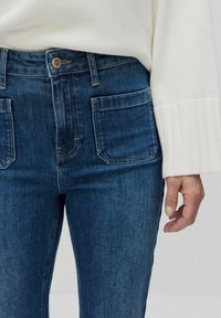 Korkeavyötär öiset siniset farkut, joissa on kaksi etutaskua ja tiukka muotoilu. Yhdistetty valkoiseen teksturoituun neuleeseen, jossa on ribbireunukset.