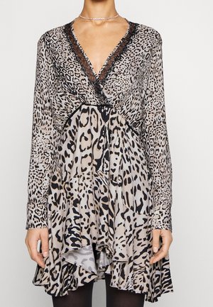 Kvinde iført en leopardmønstret slå-om-kjole med lange ærmer, sort blondetrim og flæsekant, kombineret med sorte strømpebukser og en sølvhalskæde.