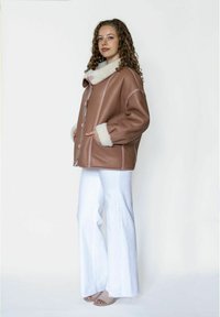 Bruine leren jas met een pluizige witte kraag, knopen aan de voorkant en twee zijzakken; gedragen over witte broek met wijde pijpen.