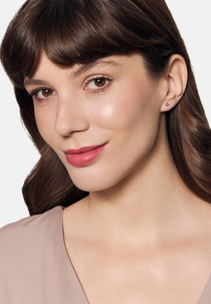 Małe kolczyki-stud z błyszczącym akcentem, zestawione z gładką, jasną bluzką. Delikatny, naturalny makijaż podkreśla neutralny wygląd.