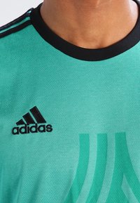 Turkusowa koszulka sportowa z czarnym kołnierzykiem. Posiada czarne logo "adidas" oraz charakterystyczne paski w jaśniejszym odcieniu turkusu na tkaninie.